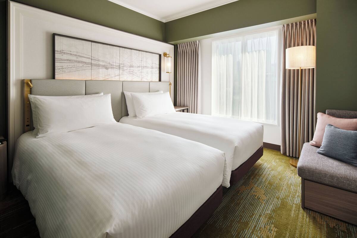 Une chambre d’hôtel avec deux lits, un canapé, une grande fenêtre avec des rideaux et une œuvre d’art accrochée au mur.