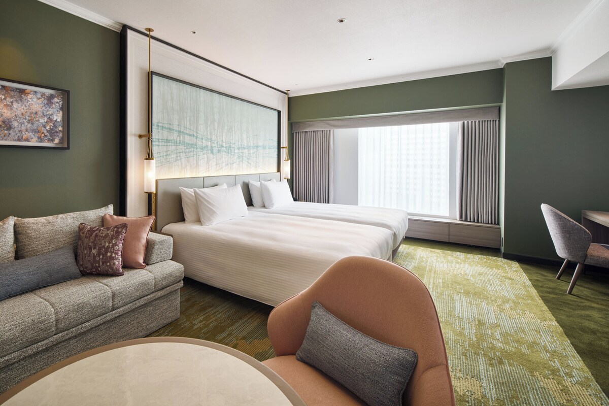 A hotel room with a large bed, a sofa, a chair, and a view of the city through the window.