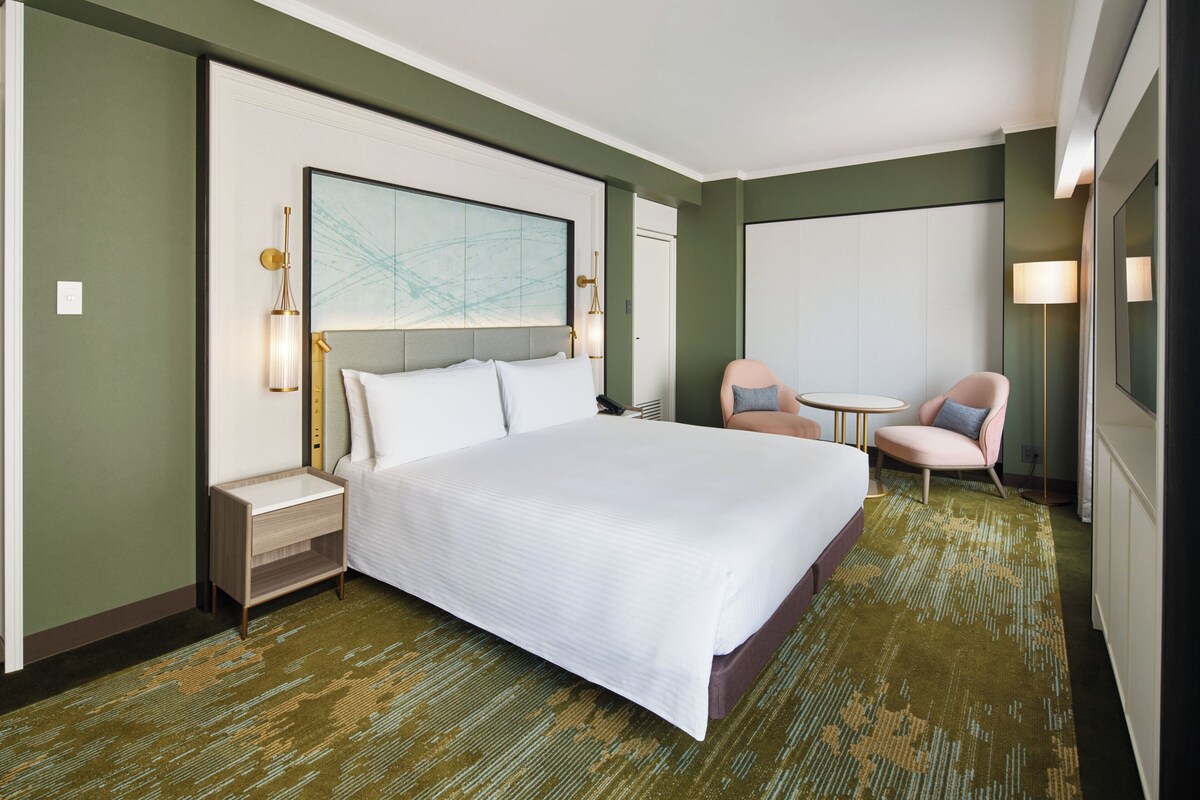 A hotel room with a large bed, a nightstand, a chair, a small table, and a wall-mounted lamp.