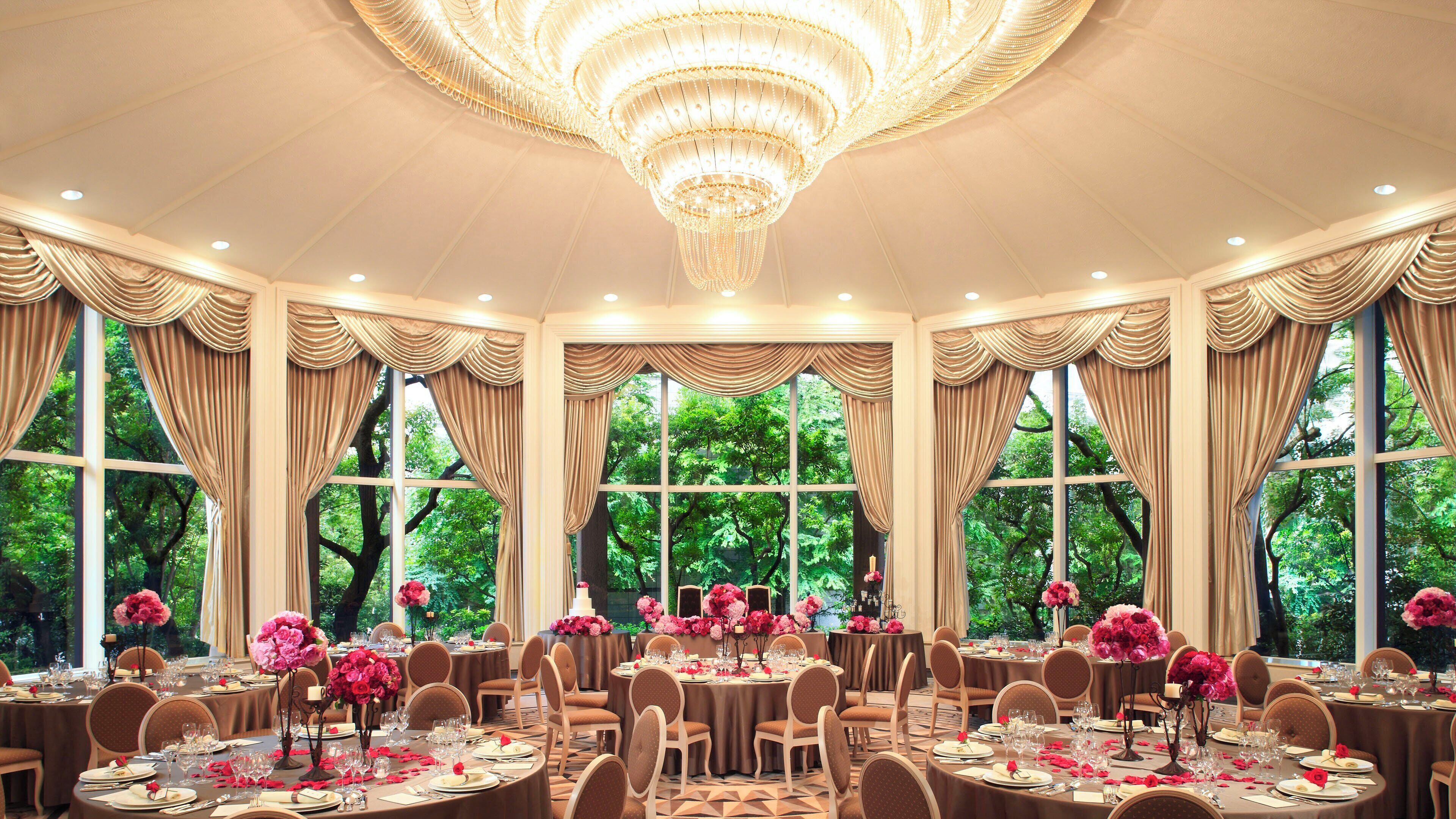 A formal dining room with round tables, chandeliers, and large windows with curtains.