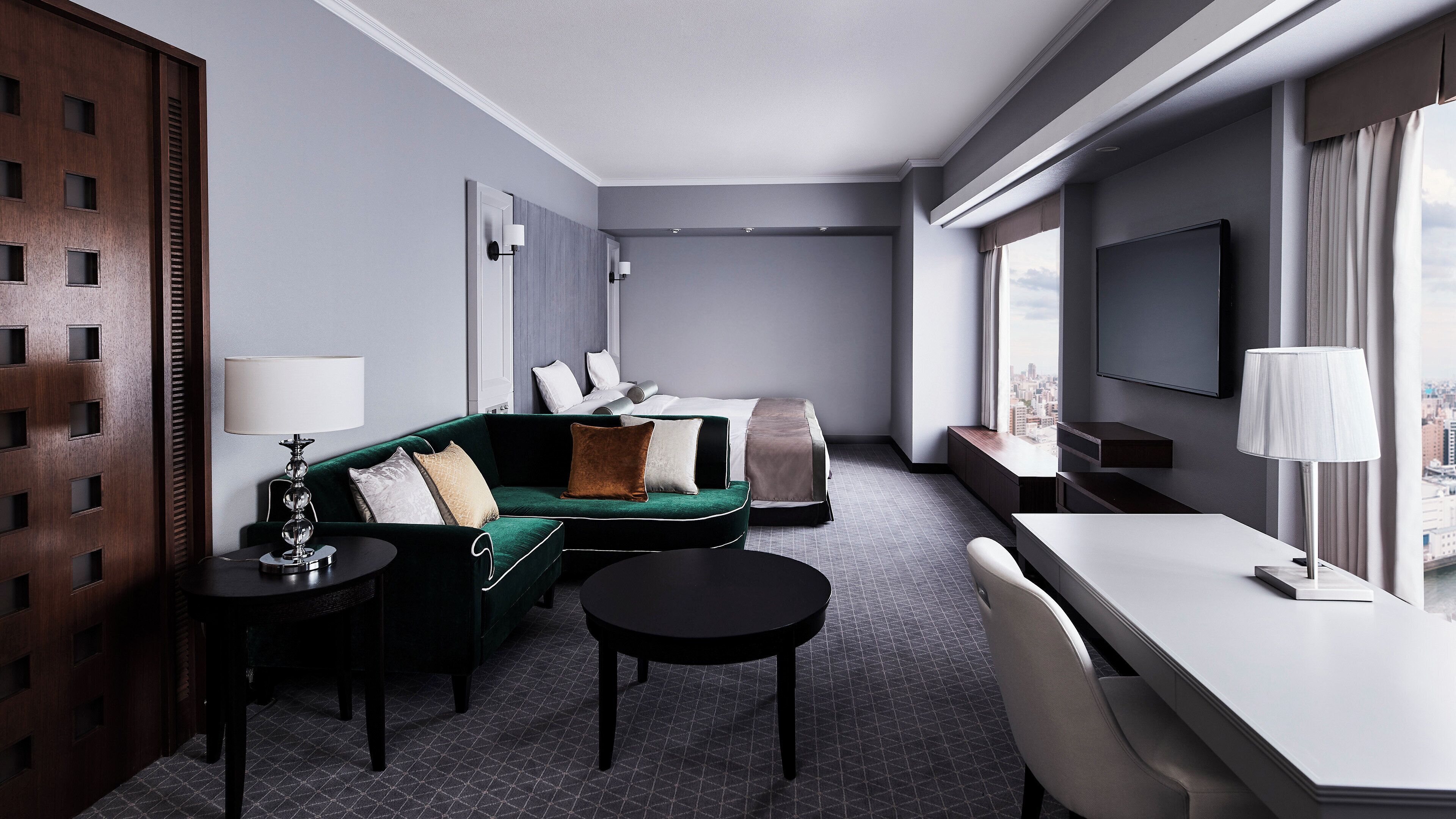A modern hotel room with a green sofa, a desk, a TV, and a city view.