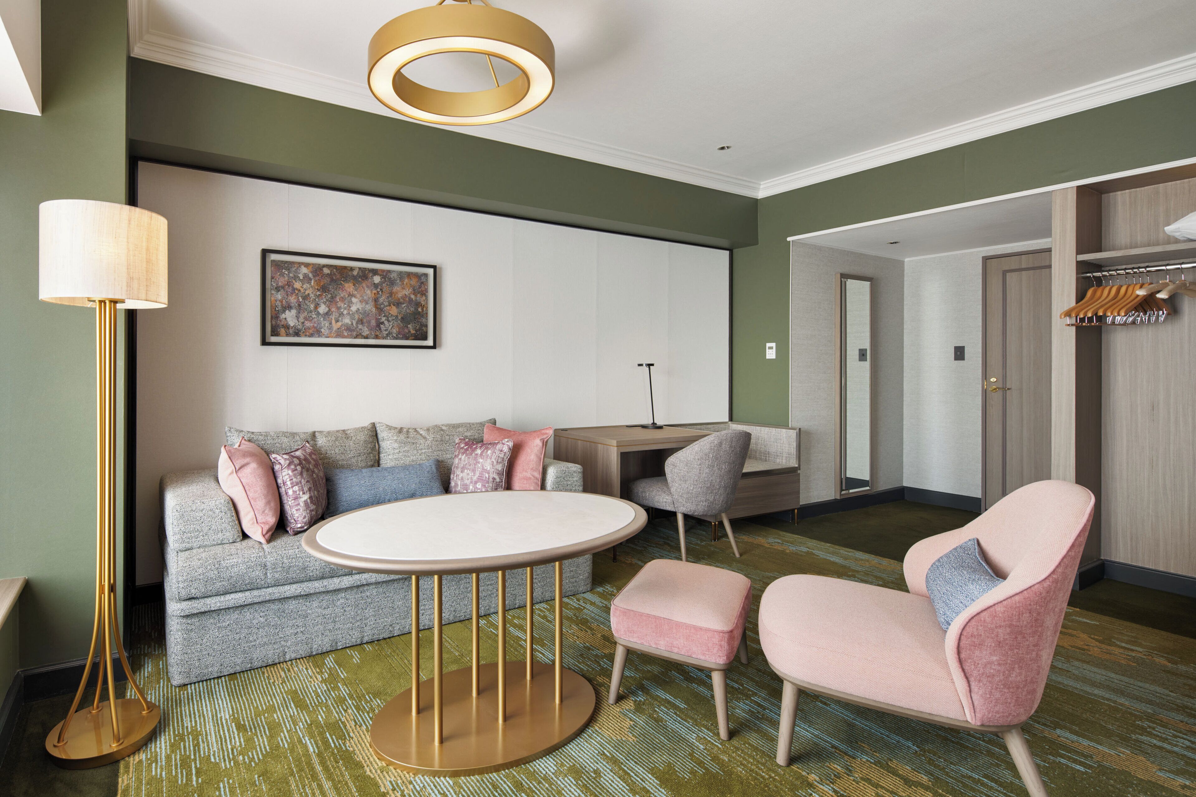 A modern hotel room with a sofa, armchair, round table, and a desk.