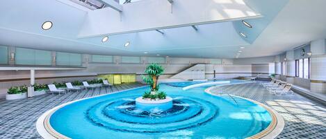 An indoor swimming pool with a central island, surrounded by lounge chairs and a tiled floor.