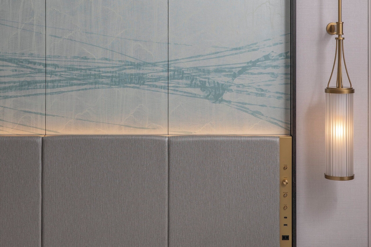 A modern hotel room with a large bed, a wall-mounted panel with a subtle design, and a classic-style wall lamp.