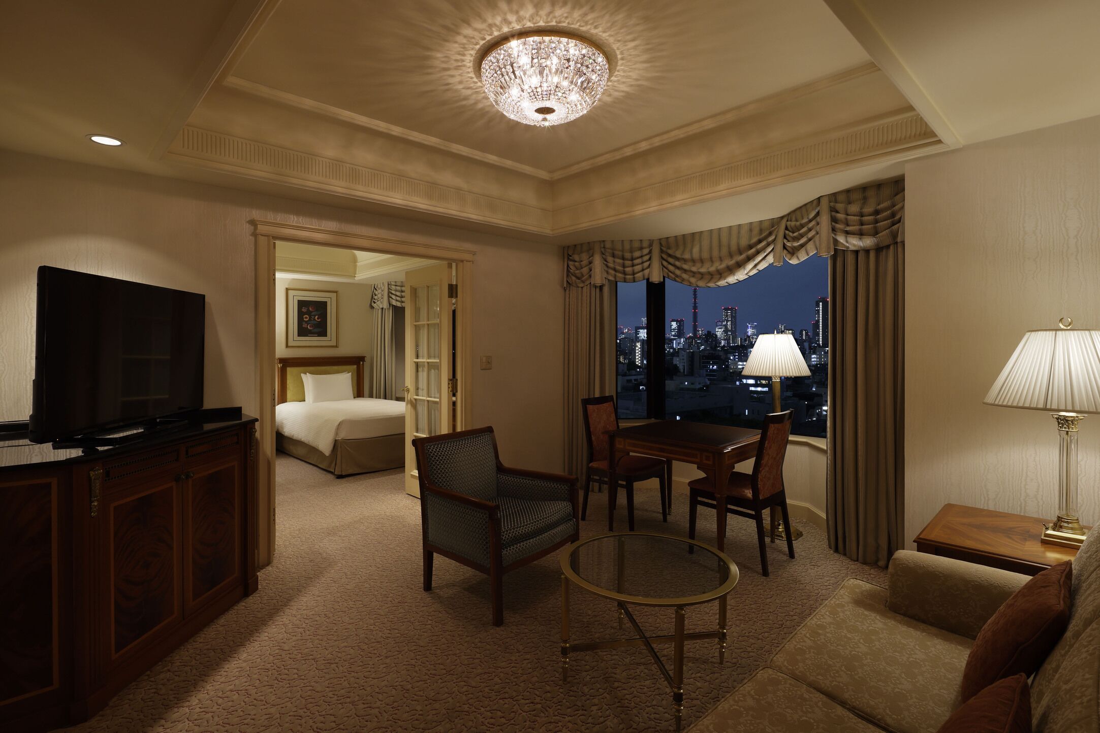 A hotel room with a television, a dining table, a sofa, a chair, a lamp, and a view of a cityscape at night.