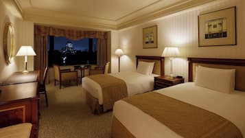 A hotel room with two beds, a desk, a chair, and a view of a cityscape at night.