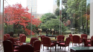 A dining area with red chairs and a table, a view of trees with red foliage, and a stone lantern in the background.