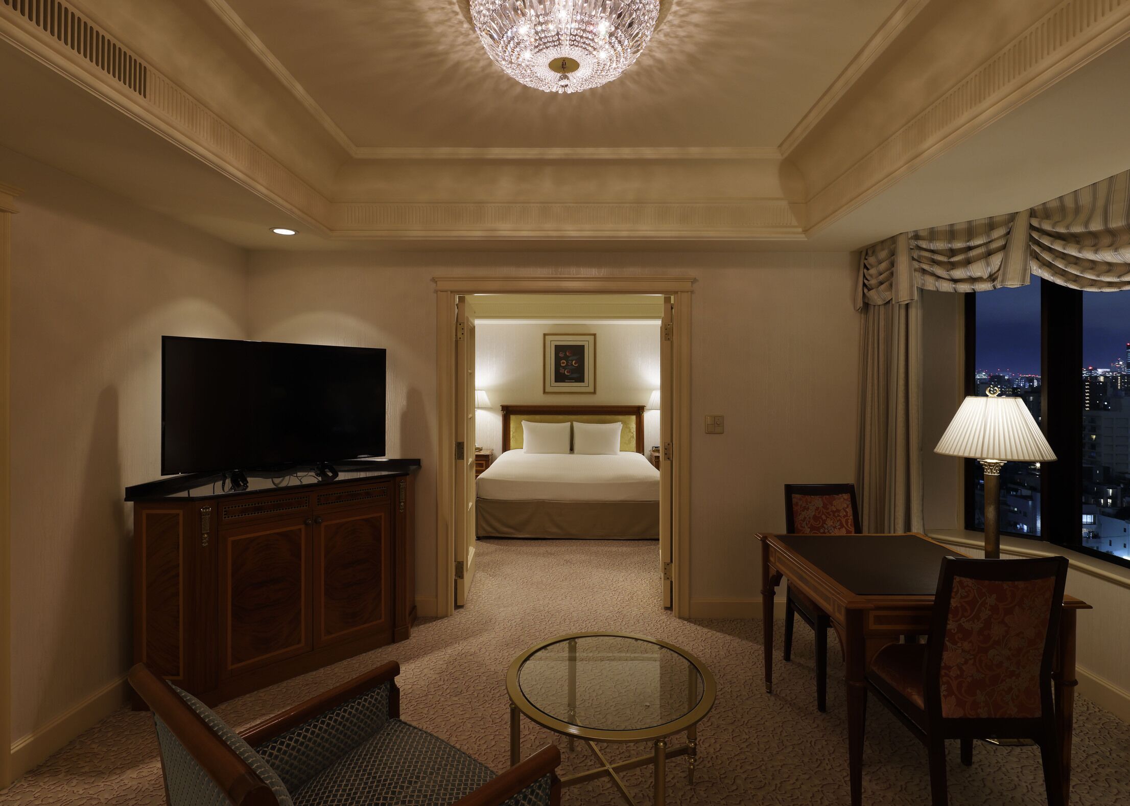 A hotel room with a television, a bed, a desk with a chair, a lamp, and a view of the city at night.