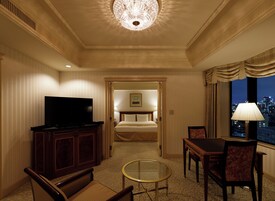 A hotel room with a television, a dining table with chairs, a bed with a headboard, and a view of the cityscape through a window.