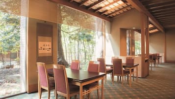 Un restaurante con mesas y sillas de madera, ventanas amplias con persianas y vistas a árboles.
