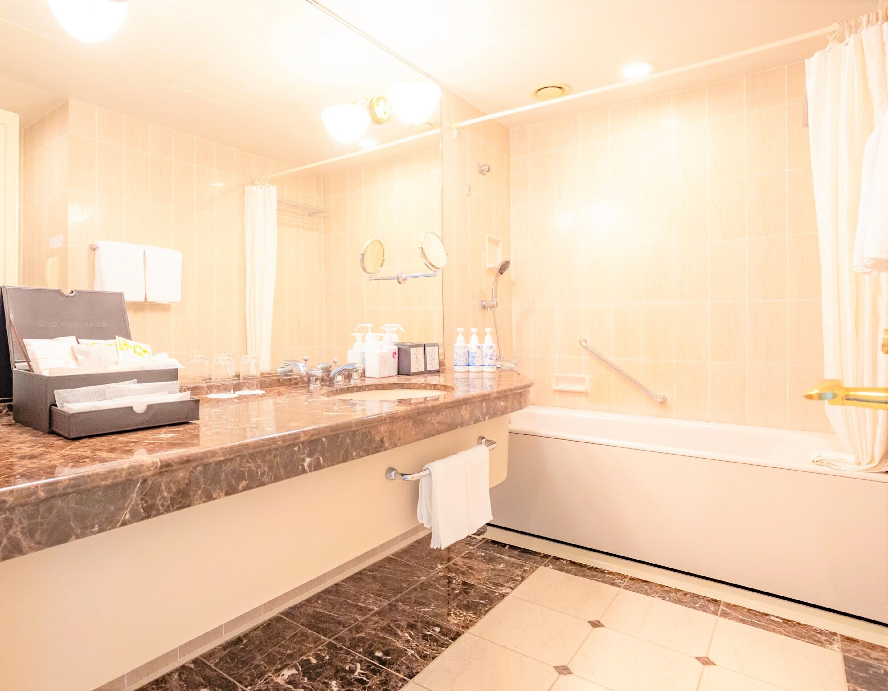 A hotel bathroom with a marble countertop, a sink, a mirror, a shower, and a bathtub. There is a towel rack and a box of toiletries on the counter.
