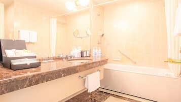 A hotel bathroom with a marble countertop, a sink, a mirror, a shower, and a bathtub. There is a towel rack and a box of toiletries on the counter.