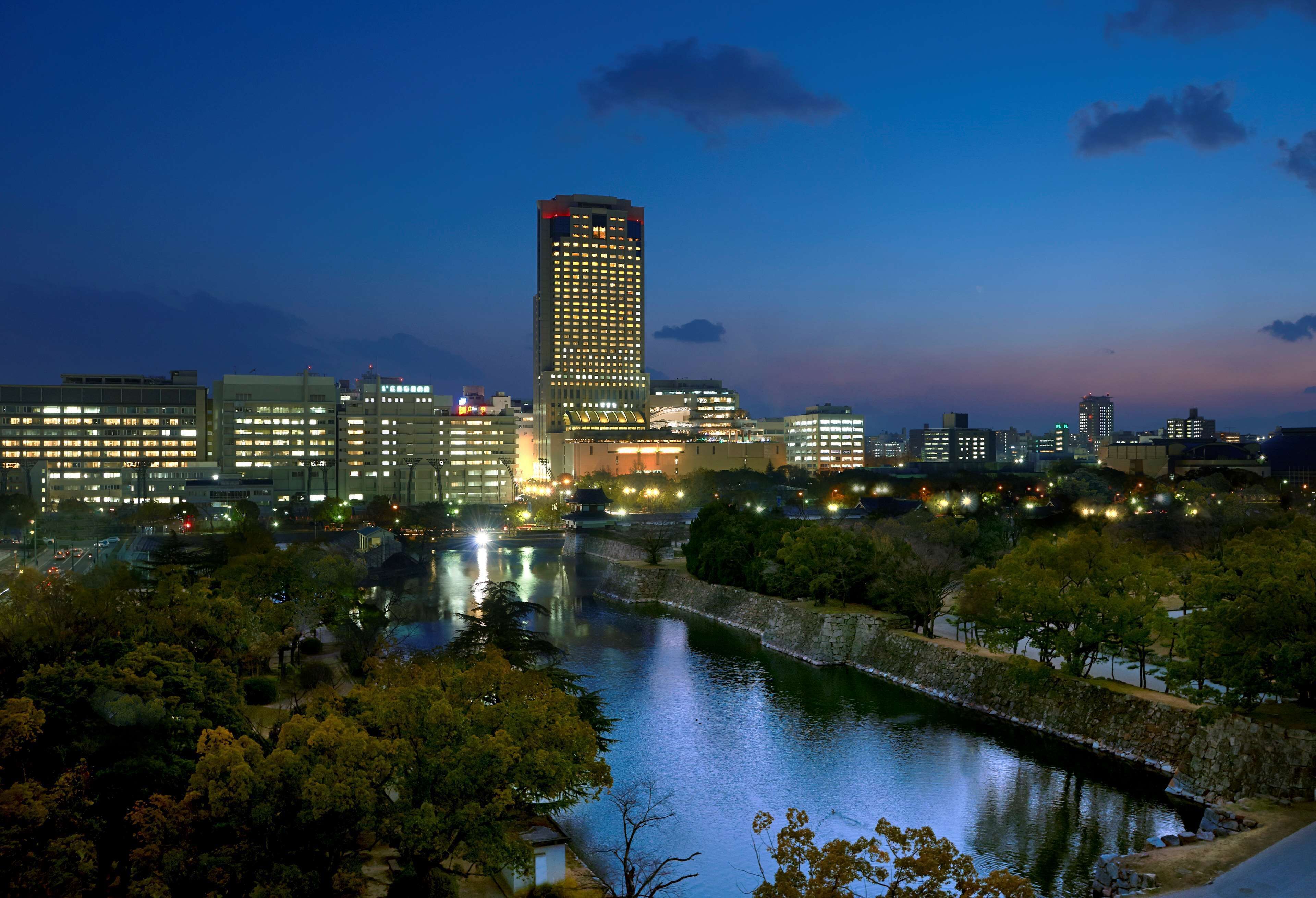 A cityscape at dusk with illuminated buildings, a river, and a park area.