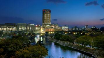 A cityscape at dusk with illuminated buildings, a river, and a park area.