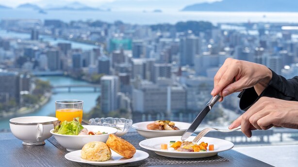 A dining table with various dishes and a glass of orange juice, overlooking a cityscape with a river and mountains. - RIHGA Royal Hotel Hiroshima (Hiroshima)