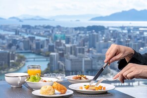 A dining table with various dishes and a glass of orange juice, overlooking a cityscape with a river and mountains. - RIHGA Royal Hotel Hiroshima (Hiroshima)