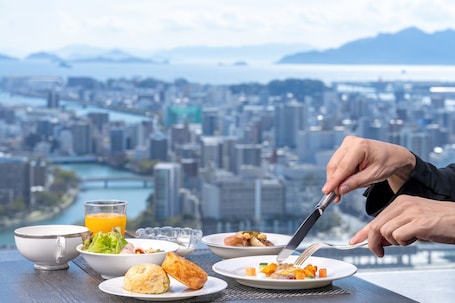 A dining table with various dishes and a glass of orange juice, overlooking a cityscape with a river and mountains.