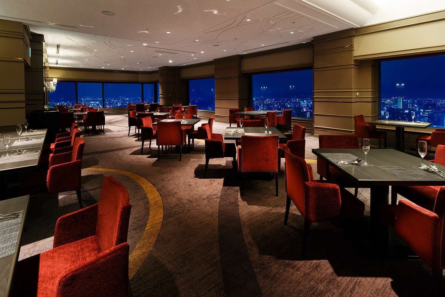 A hotel dining area with multiple tables and chairs, large windows offering a city view, and a carpeted floor.