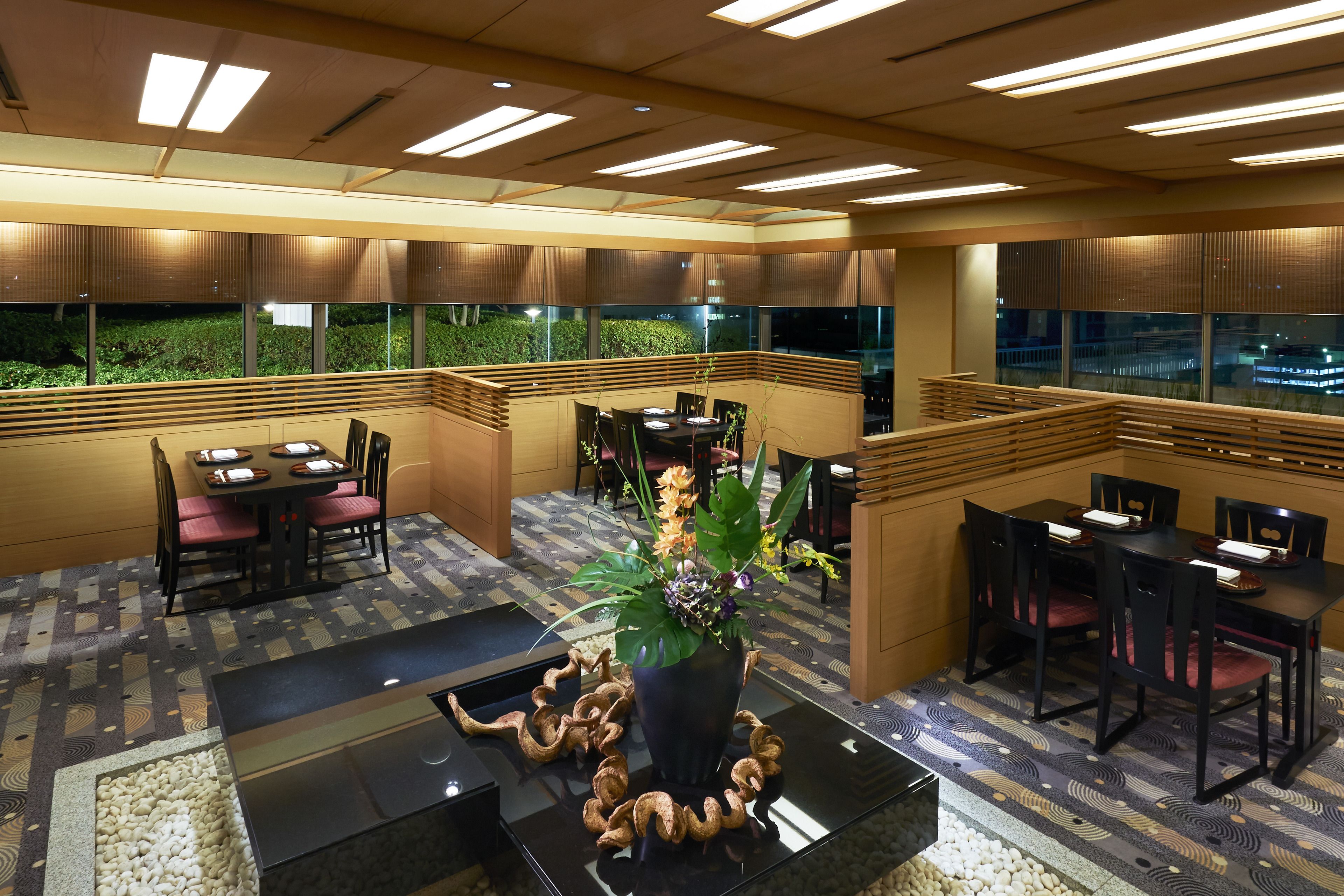 Un comedor moderno con mesas y sillas, una planta decorativa en el centro y vistas amplias a través de grandes ventanales.