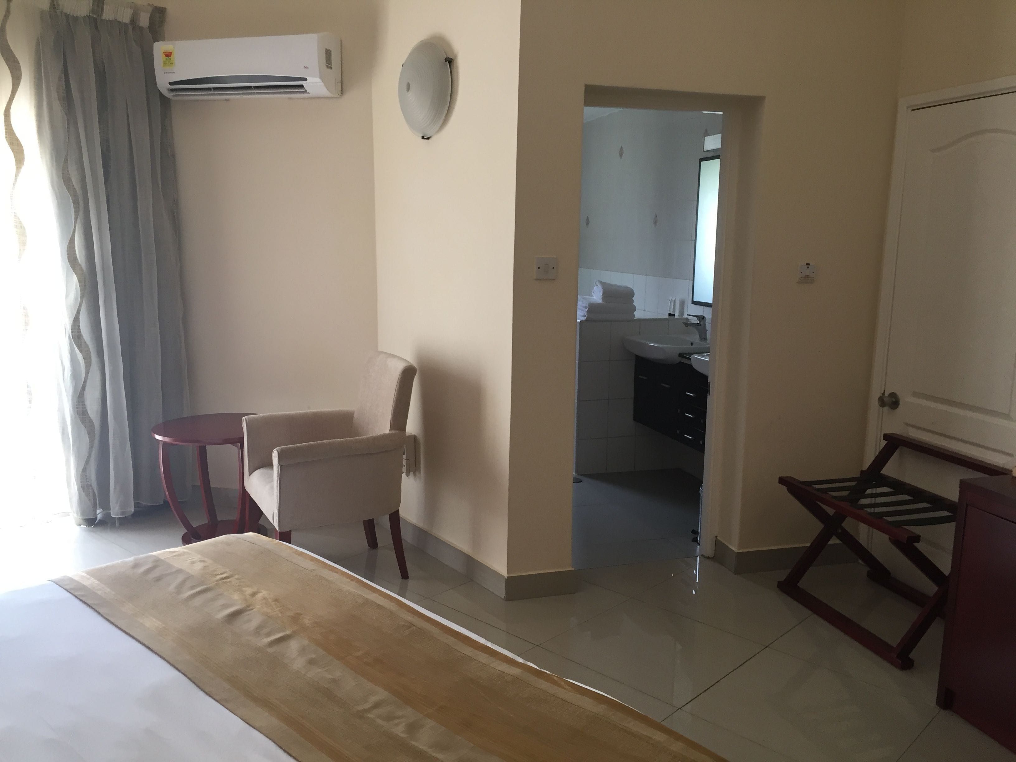 A hotel room with a bed, a small table, two chairs, an air conditioning unit, a bathroom visible through an open door, and a coat rack.