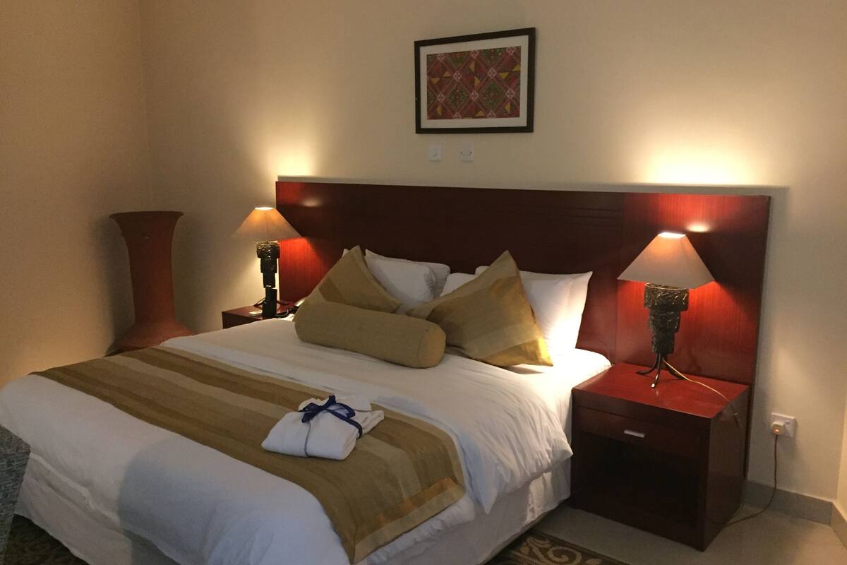 Una cama bien hecha con una colcha a rayas, dos lámparas de noche, un cabecero de madera y un cuadro enmarcado en la pared.