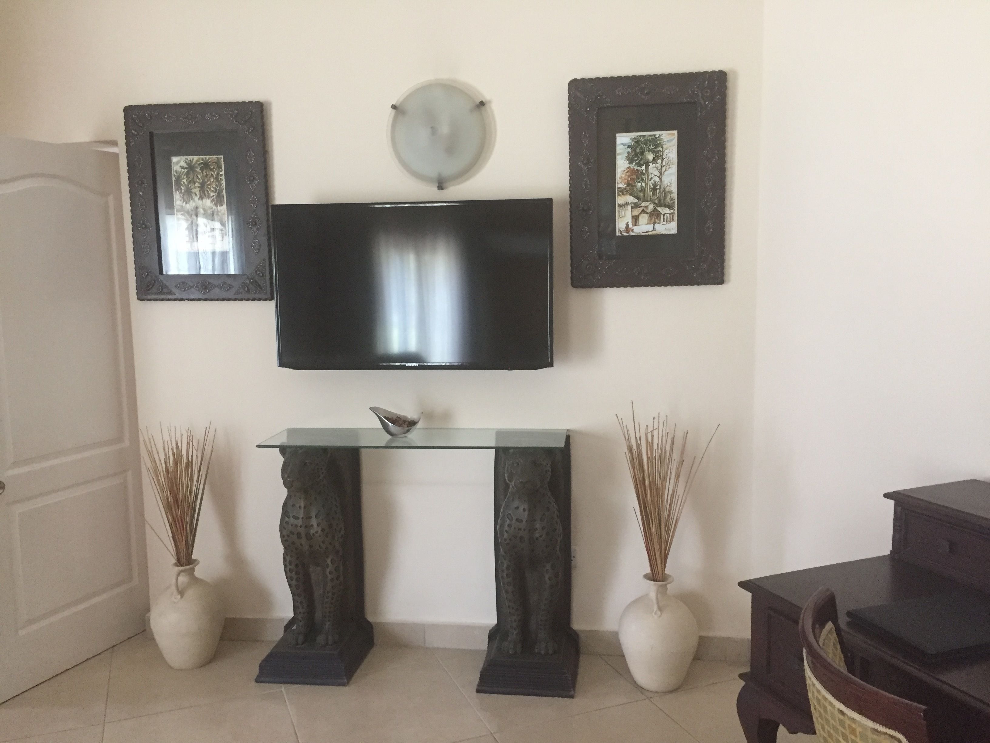 A living room with a flat-screen TV mounted on the wall, flanked by two decorative vases with tall grasses, a small bowl on a glass shelf, and a dark wooden cabinet to the right.