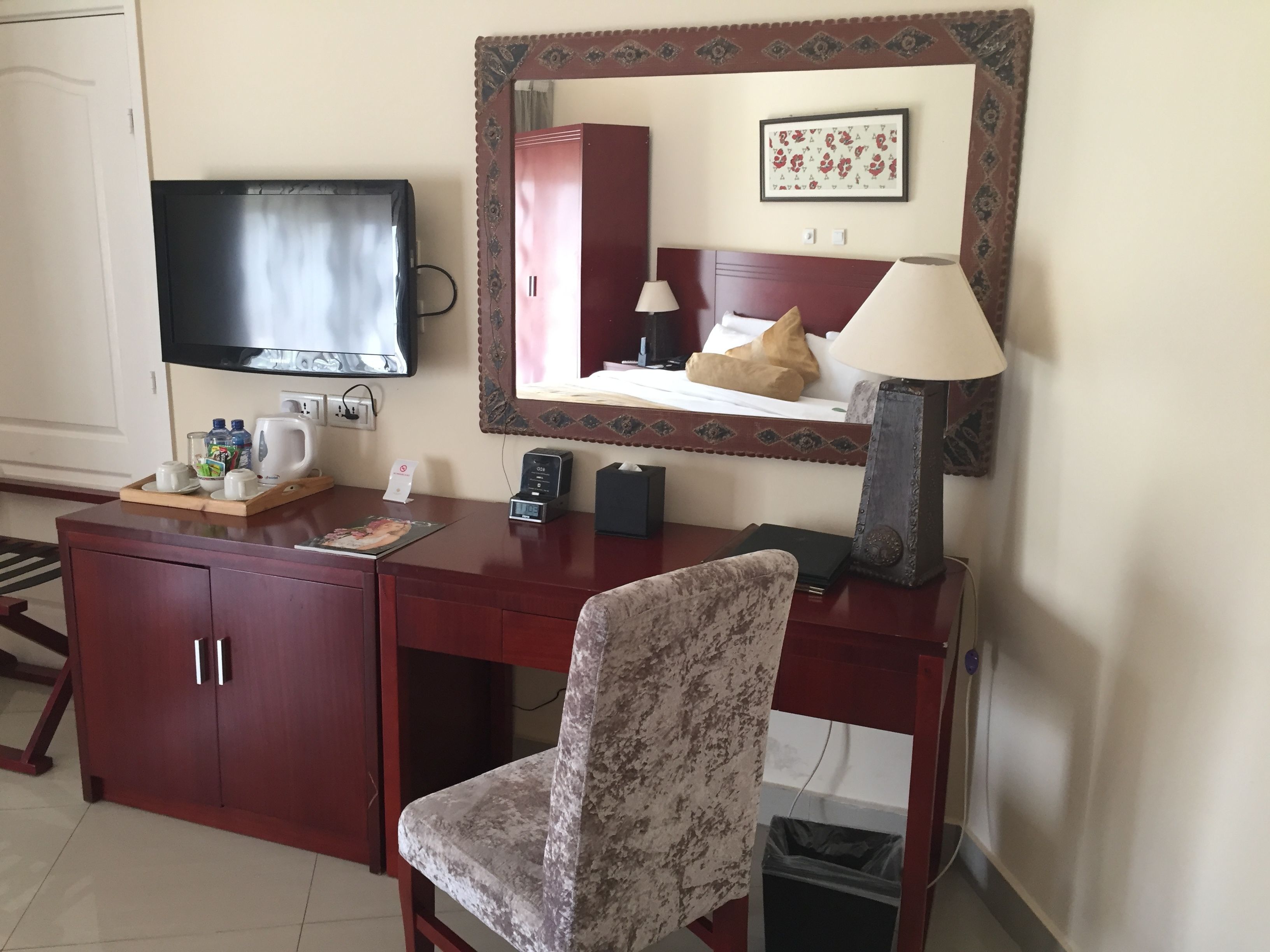 Hotelkamer met een houten bureau, een televisie, een bed met een rode hoofdeinde, een spiegel met decoratieve lijst en een kleine prullenbak.