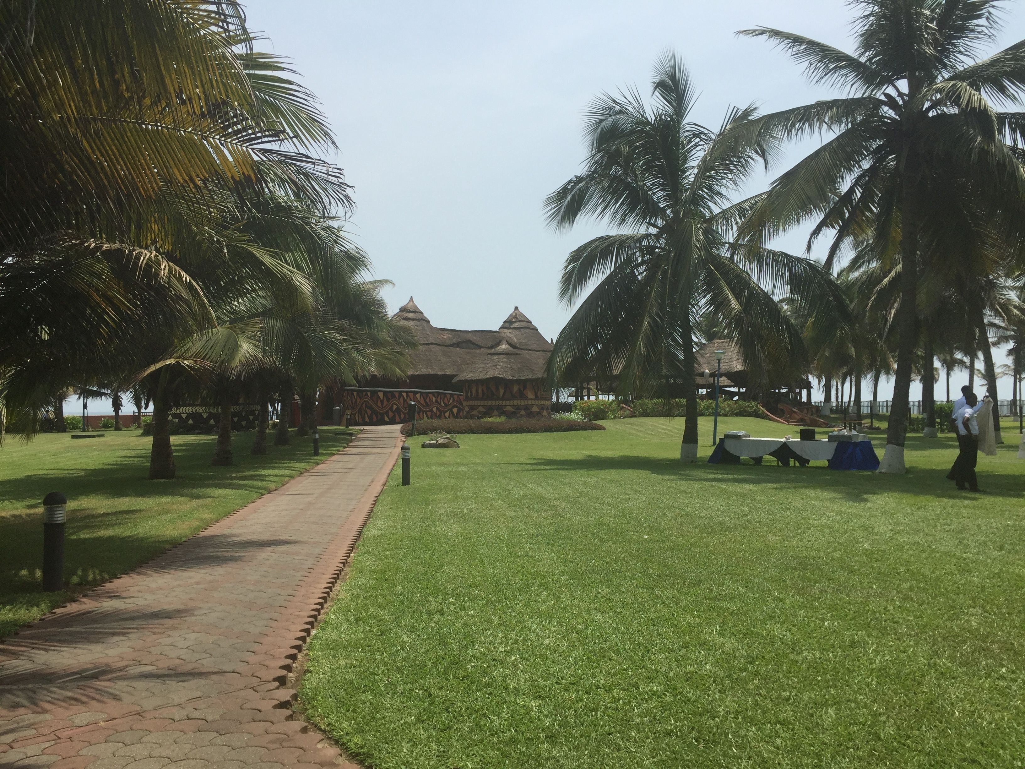 A pathway leading to a building with a thatched roof, surrounded by palm trees and a well-maintained lawn.