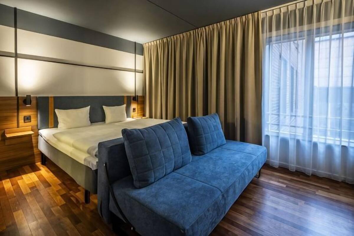 Habitación de hotel con cama, sofá azul, suelo de madera, ventana grande con cortinas e iluminación en la pared.