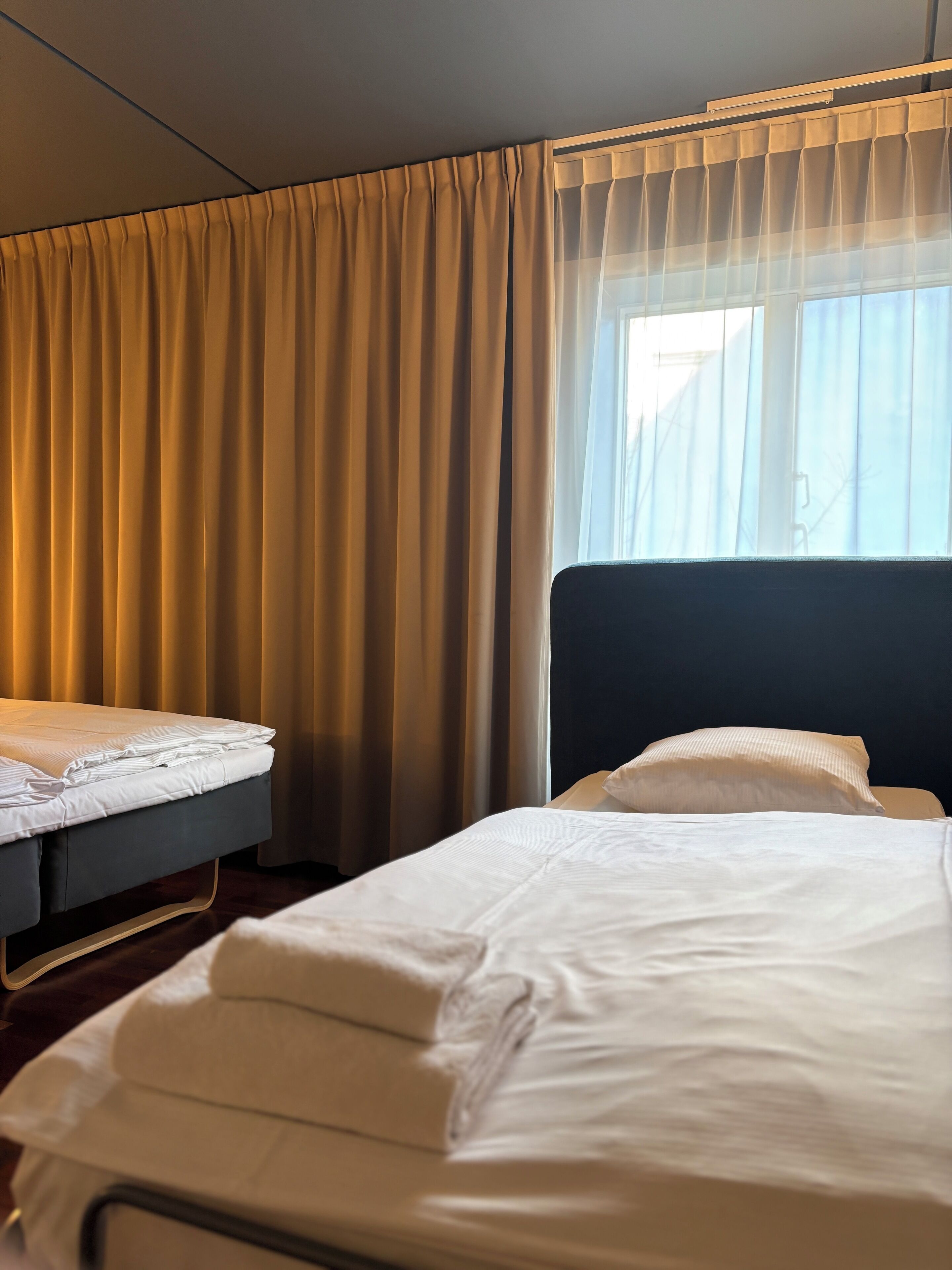 A hotel room with two beds, one with a dark headboard and the other with a white headboard. The room has beige curtains and a window with sheer white curtains.