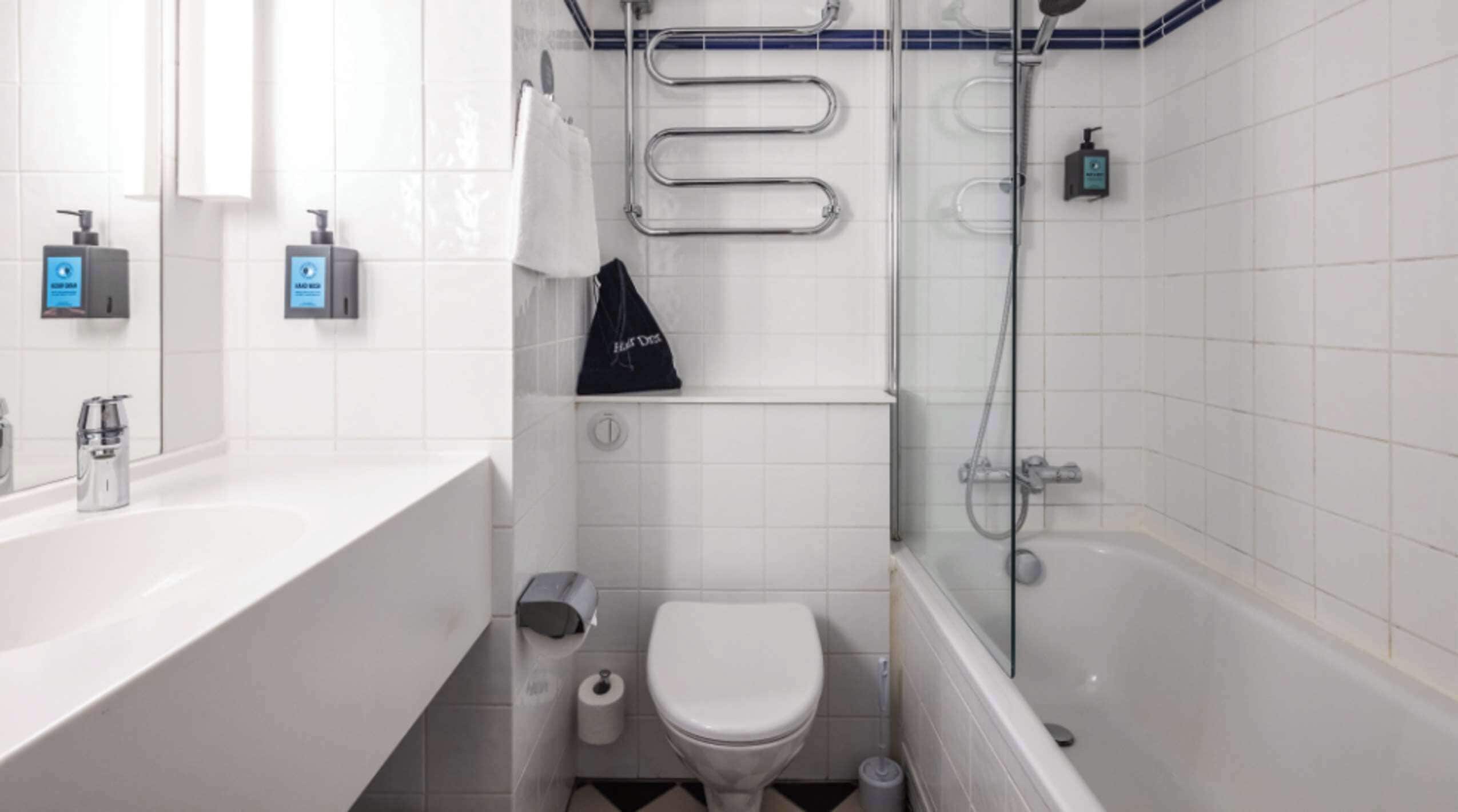 Een kleine badkamer met een wit wastafel, toilet en douche. Er zijn twee zeepdispensers aan de muur, een handdoek op een rek en een afvalbak.