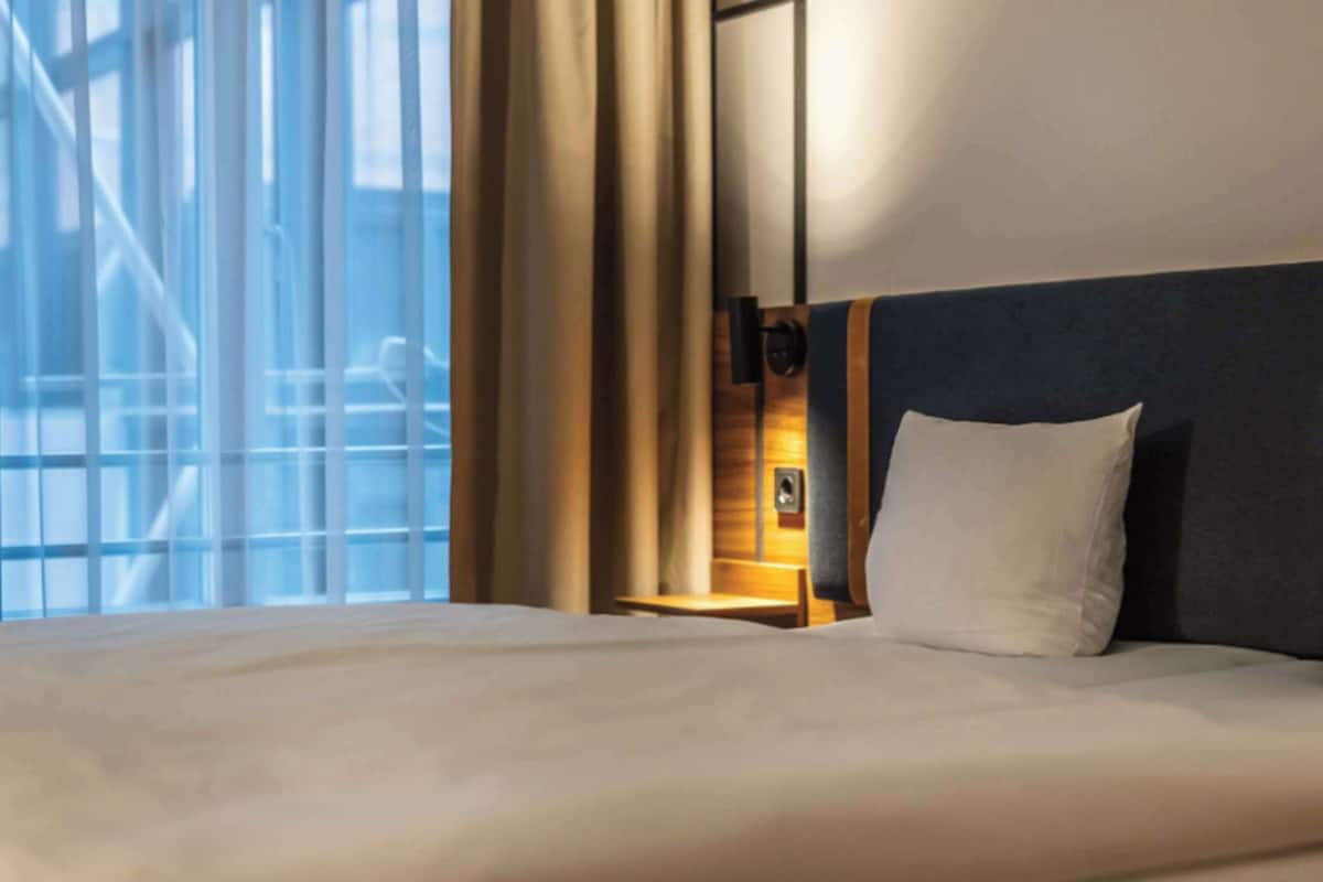 Habitación de hotel con una cama grande, una mesita de noche, una lámpara de pared y una ventana con cortinas.