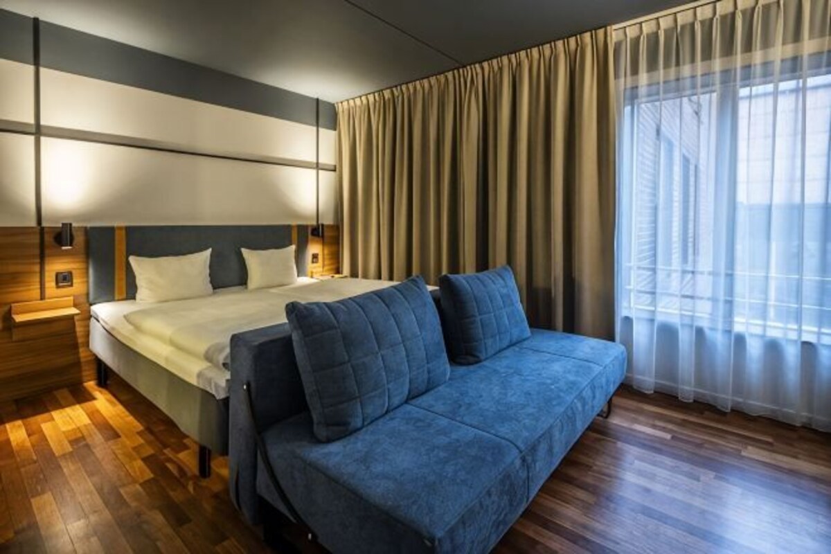 Une chambre d’hôtel avec un lit, un canapé bleu, un plancher en bois, une grande fenêtre avec des rideaux et des luminaires fixés au mur.