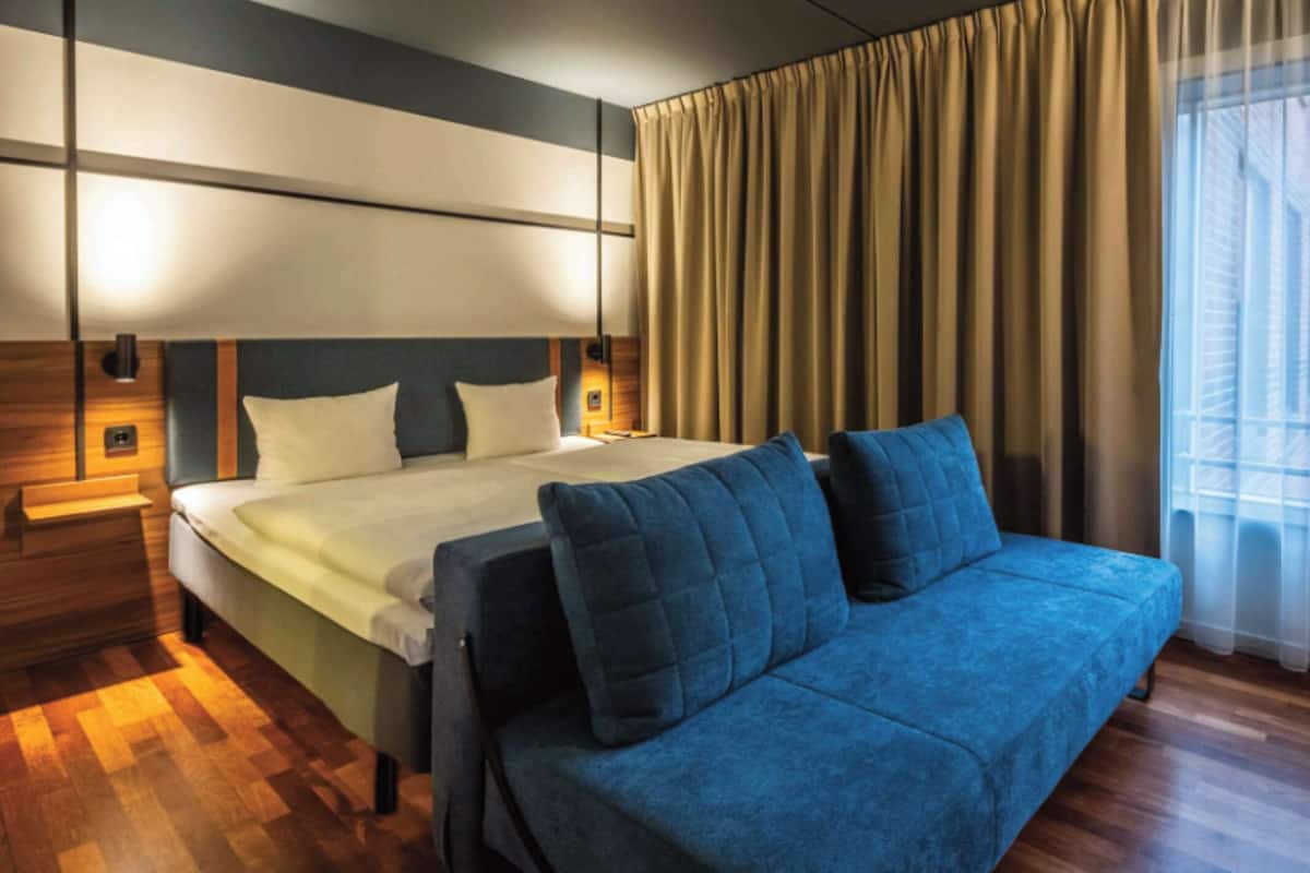 Una habitación de hotel moderna con una cama grande, un sofá azul, suelo de madera y un amplio ventanal con cortinas.