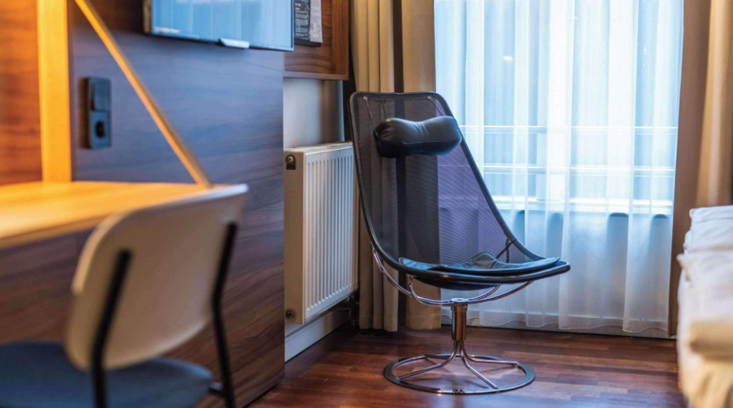 A modern hotel room with a wooden floor, a black mesh chair, a white chair, a wooden desk, and a television mounted on the wall.