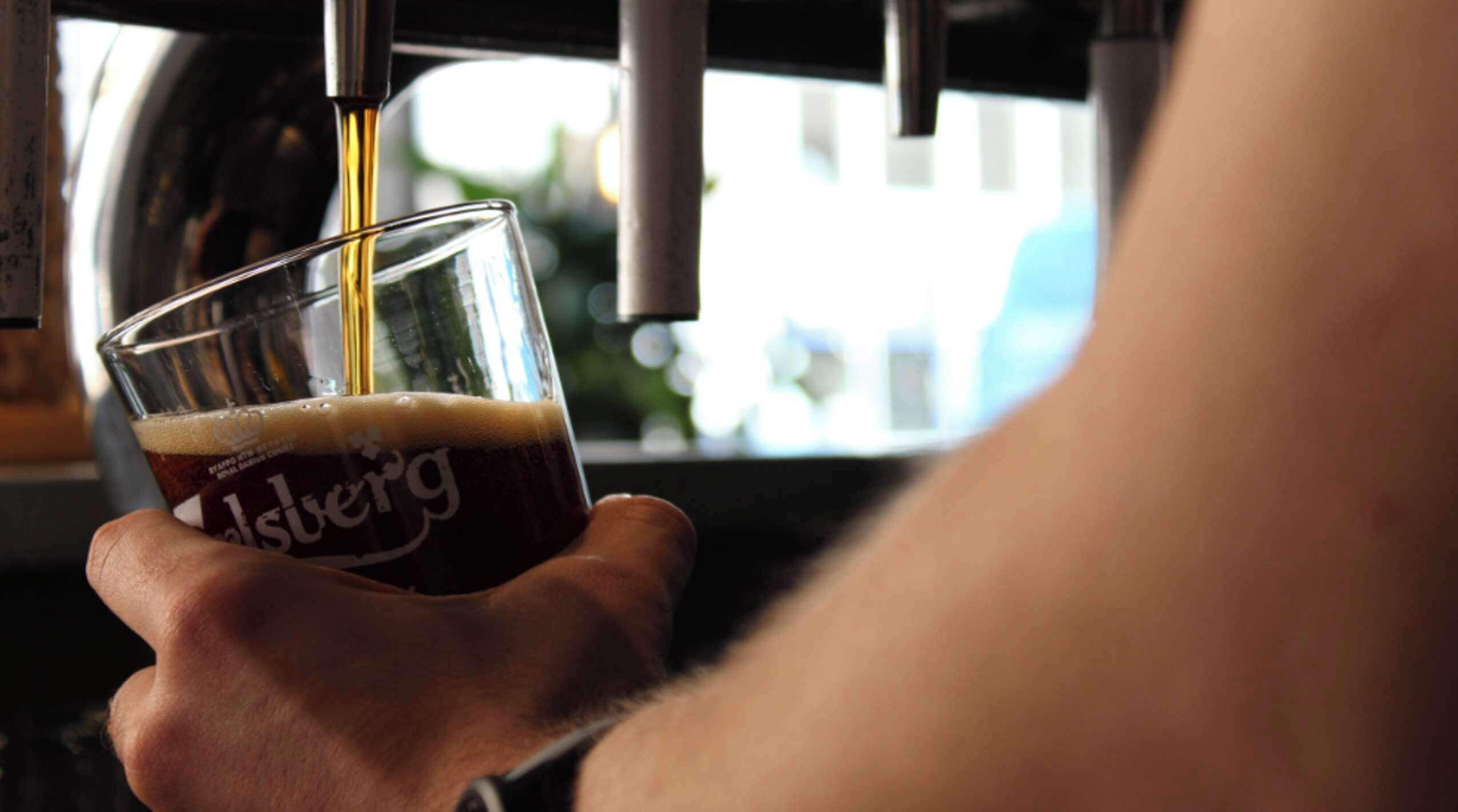 A hand holding a glass with a dark liquid, possibly beer, with a label that reads "Alsterberg."