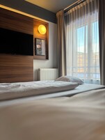 A hotel room with a bed, a mounted TV, a wall-mounted light, and a window with curtains.