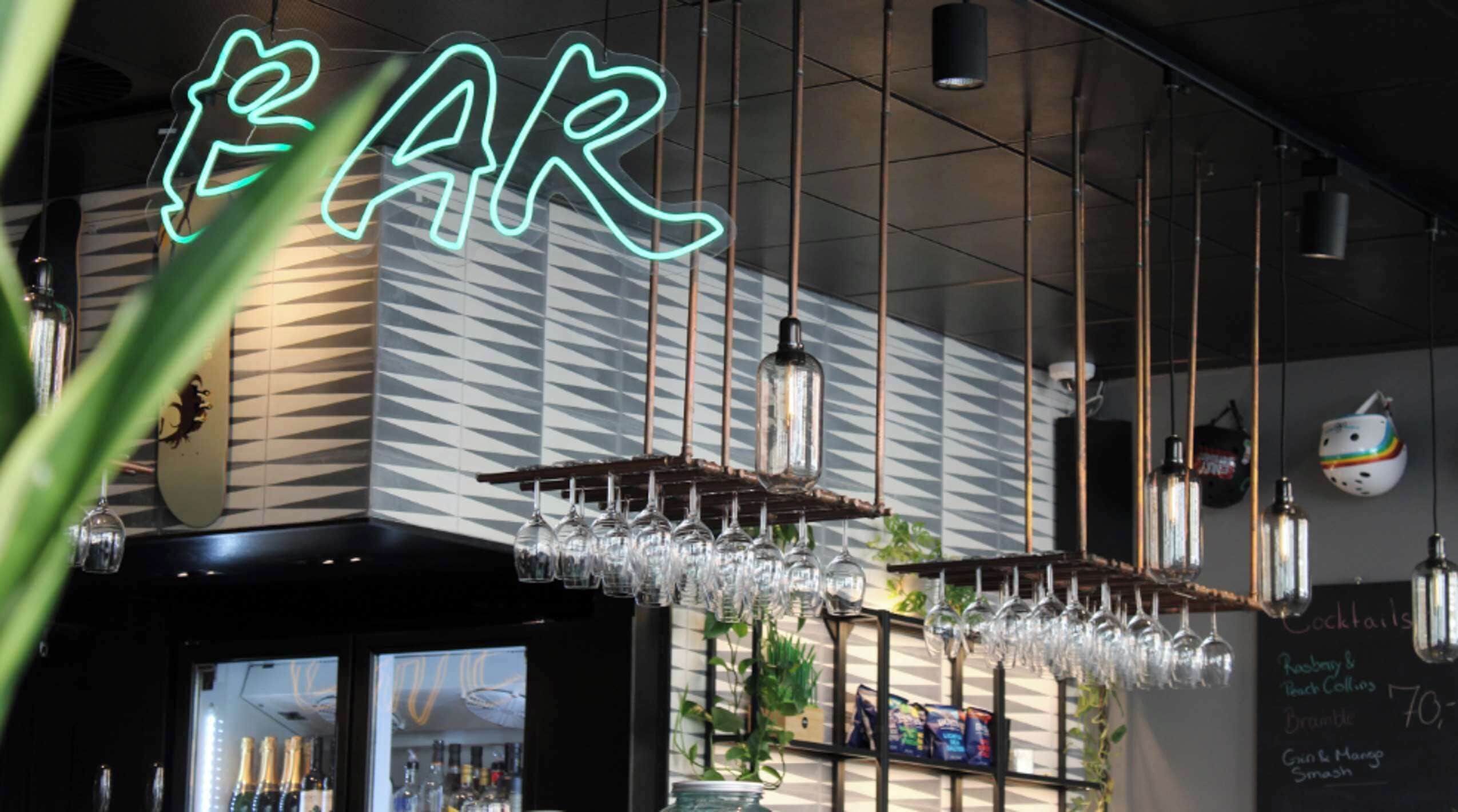 A bar area with a neon sign, hanging light fixtures, and a chalkboard menu listing cocktails.