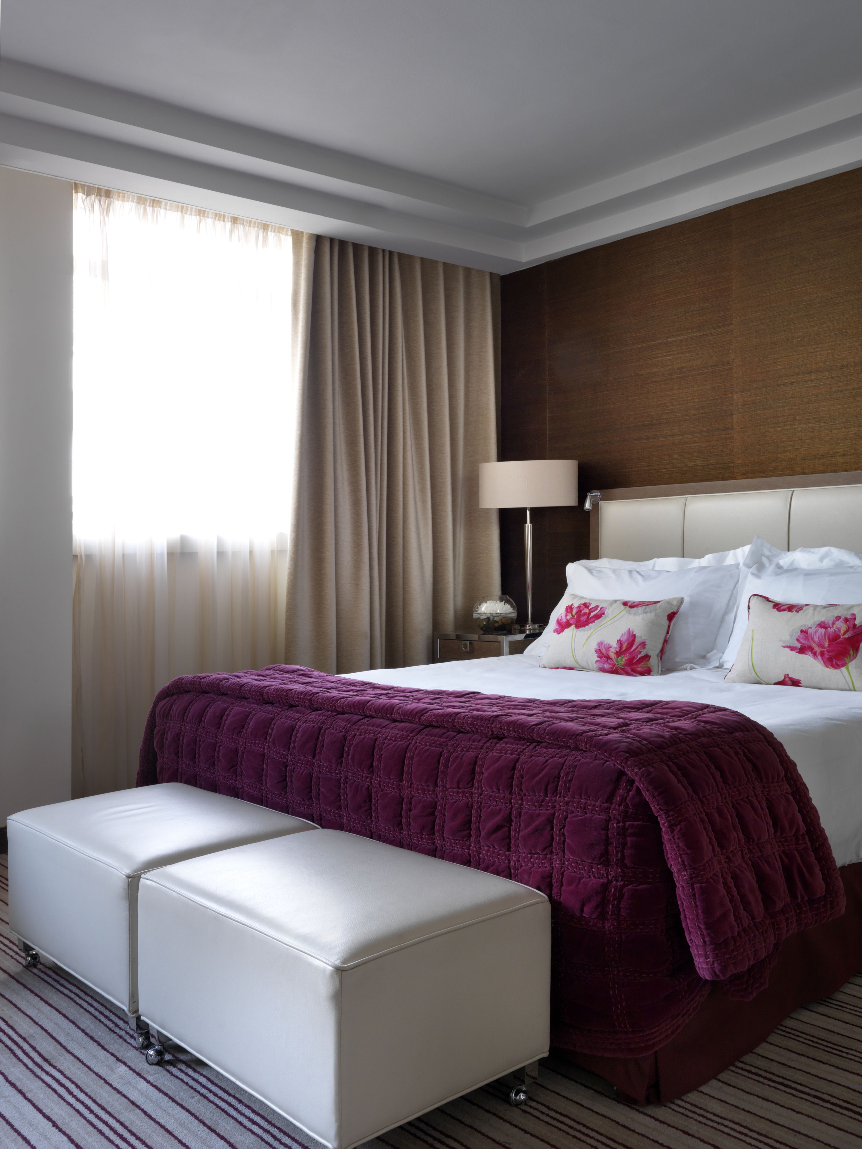 A hotel room with a large bed, a bedside table with a lamp, a window with curtains, and a small white bench.