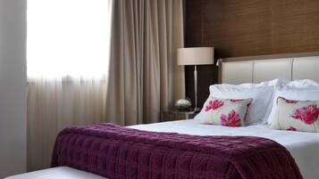 A hotel room with a large bed, a bedside table with a lamp, a window with curtains, and a small white bench.