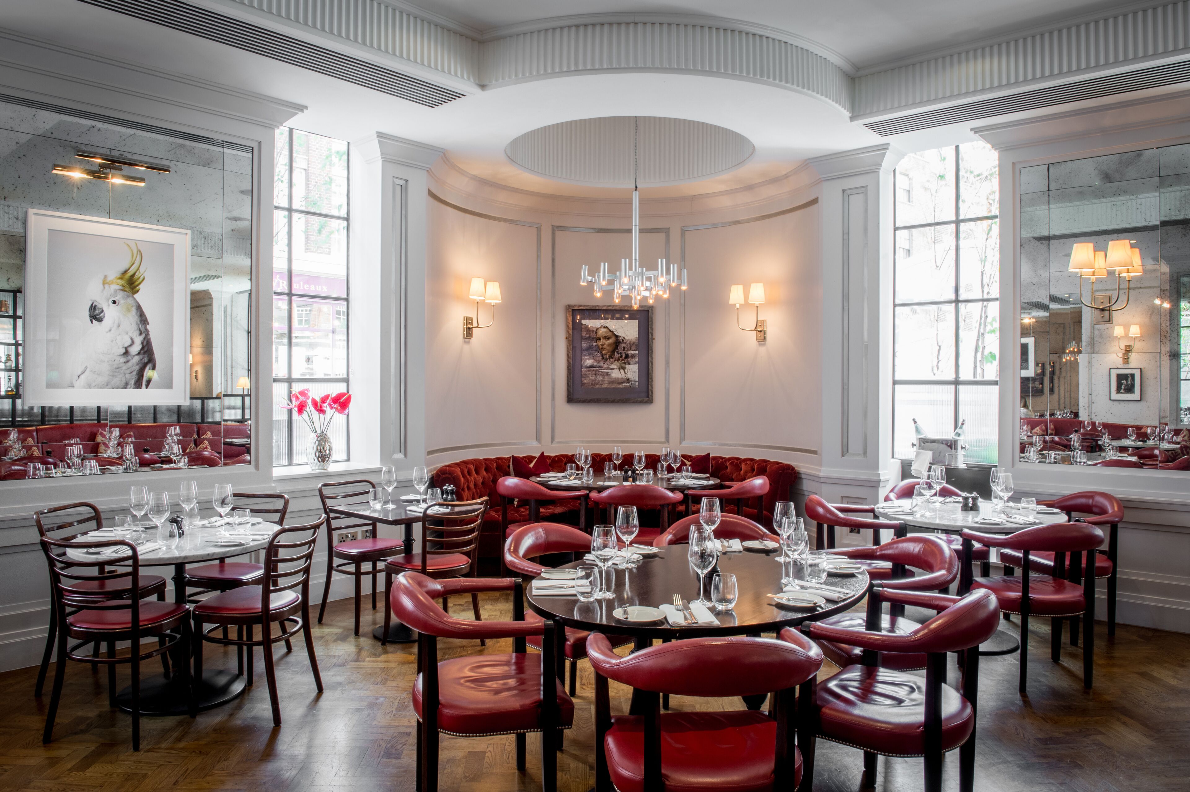 Un espace repas avec des tables rondes et des banquettes rembourrées de couleur rouge, de grandes fenêtres et des luminaires fixés au mur.