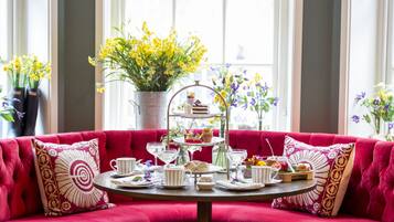 Ein rotes Sofa mit rundem Tisch, gedeckt für eine Teezeremonie, umgeben von Blumenarrangements und einem Fenster mit Vorhängen.