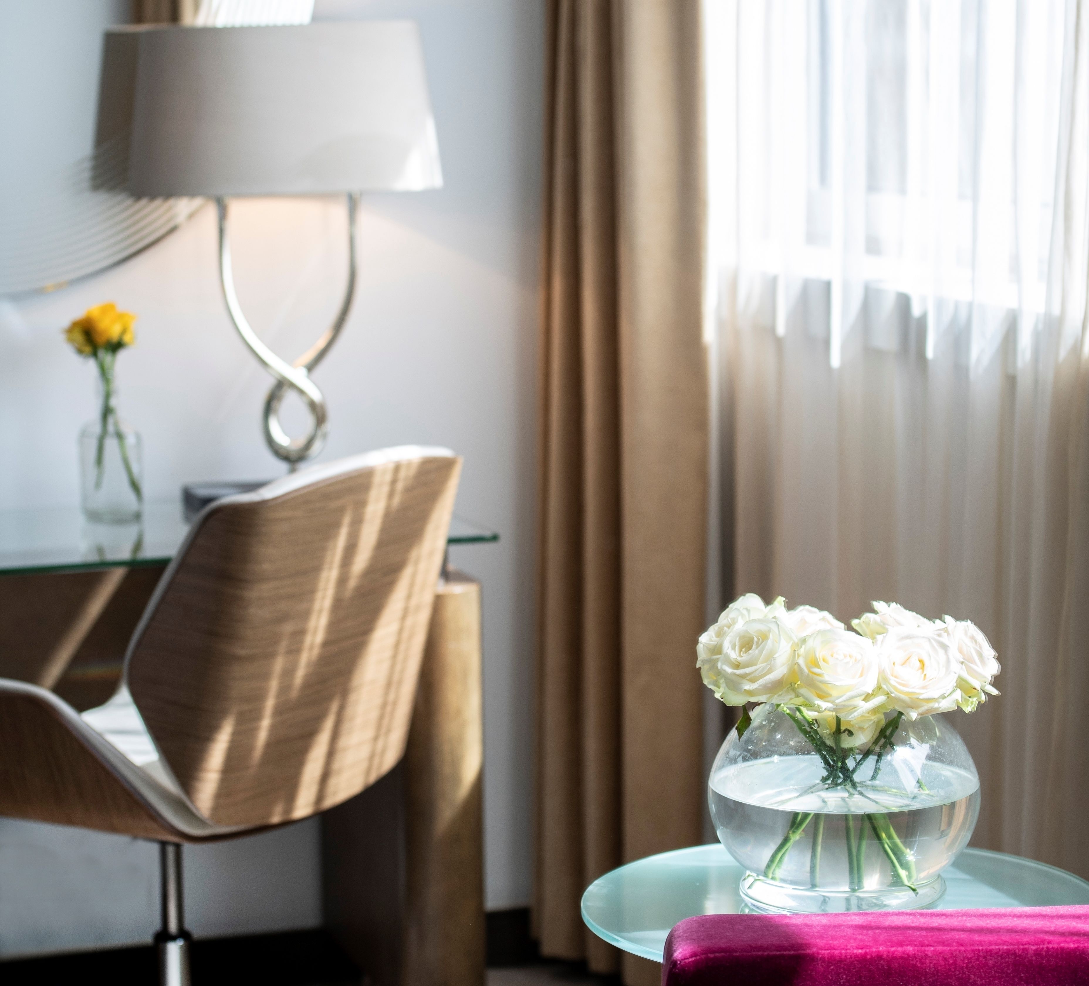A modern hotel room with a glass table holding a vase of white flowers, a desk with a lamp, and a view of the outdoors through sheer curtains.