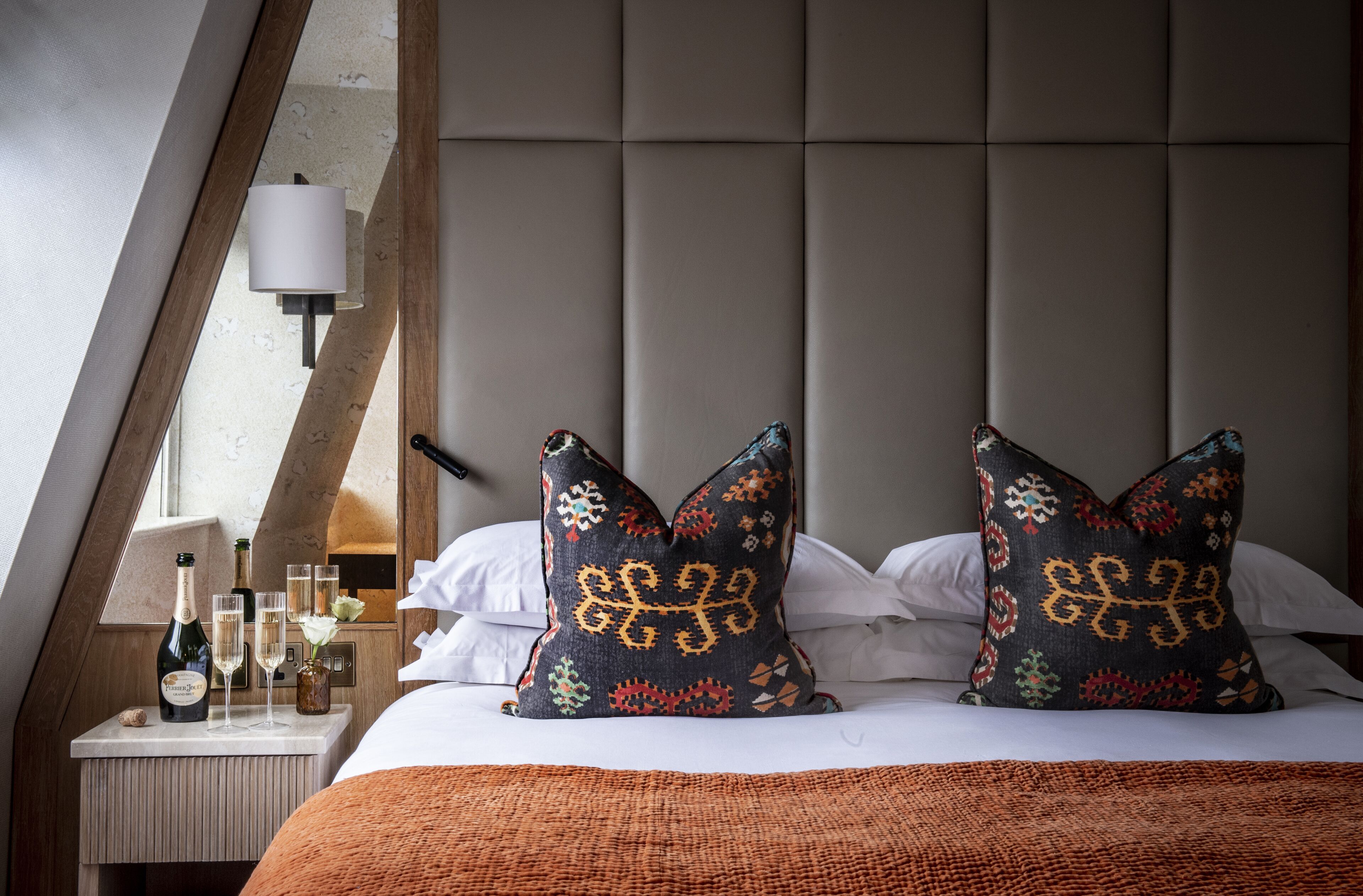 Habitación de hotel con cama, dos almohadones decorativos, mesita de noche con botella y vasos, y lámpara de pared.