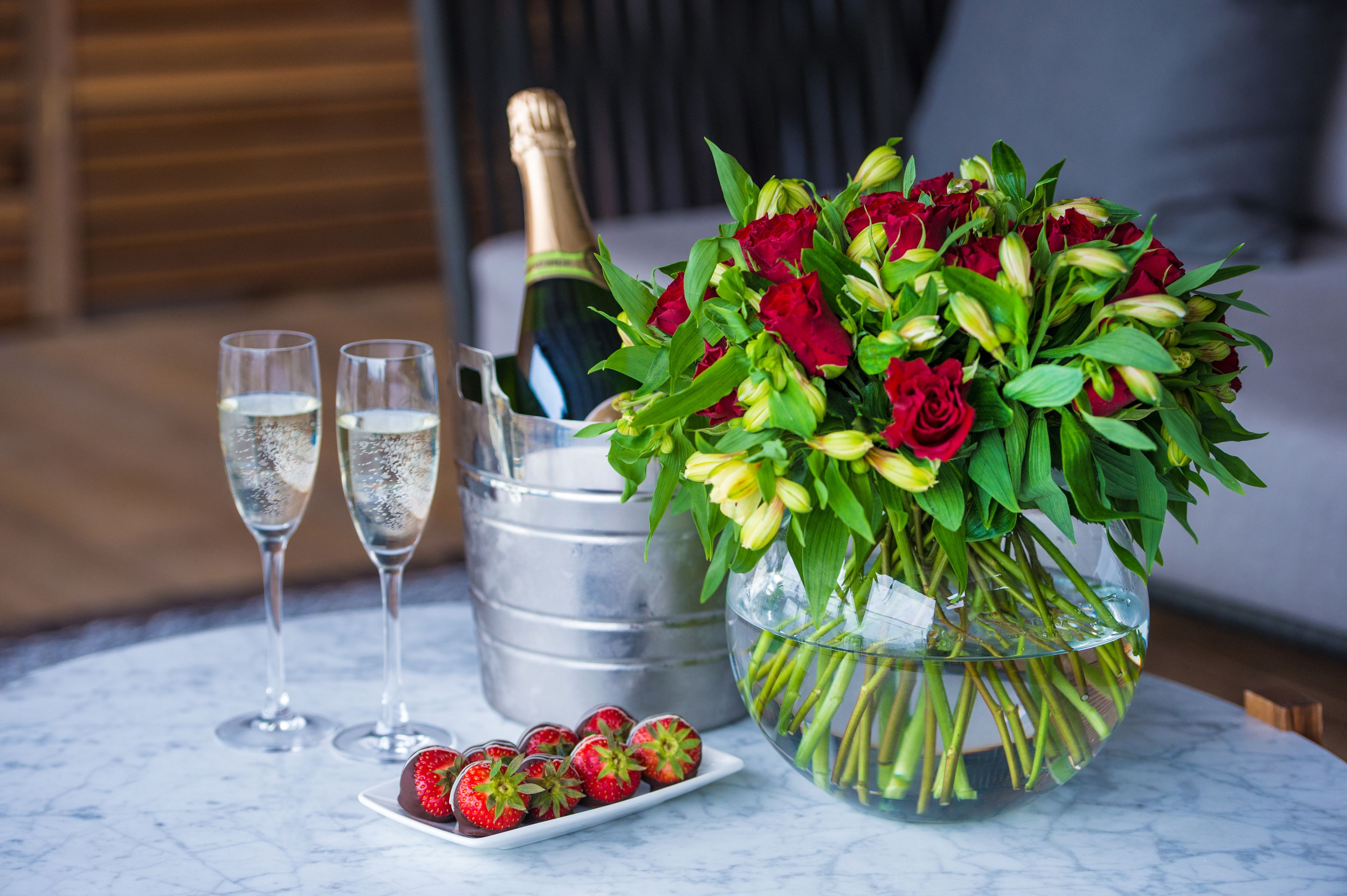 A table with a champagne bottle, two glasses, a bouquet of red roses, and a plate of strawberries.