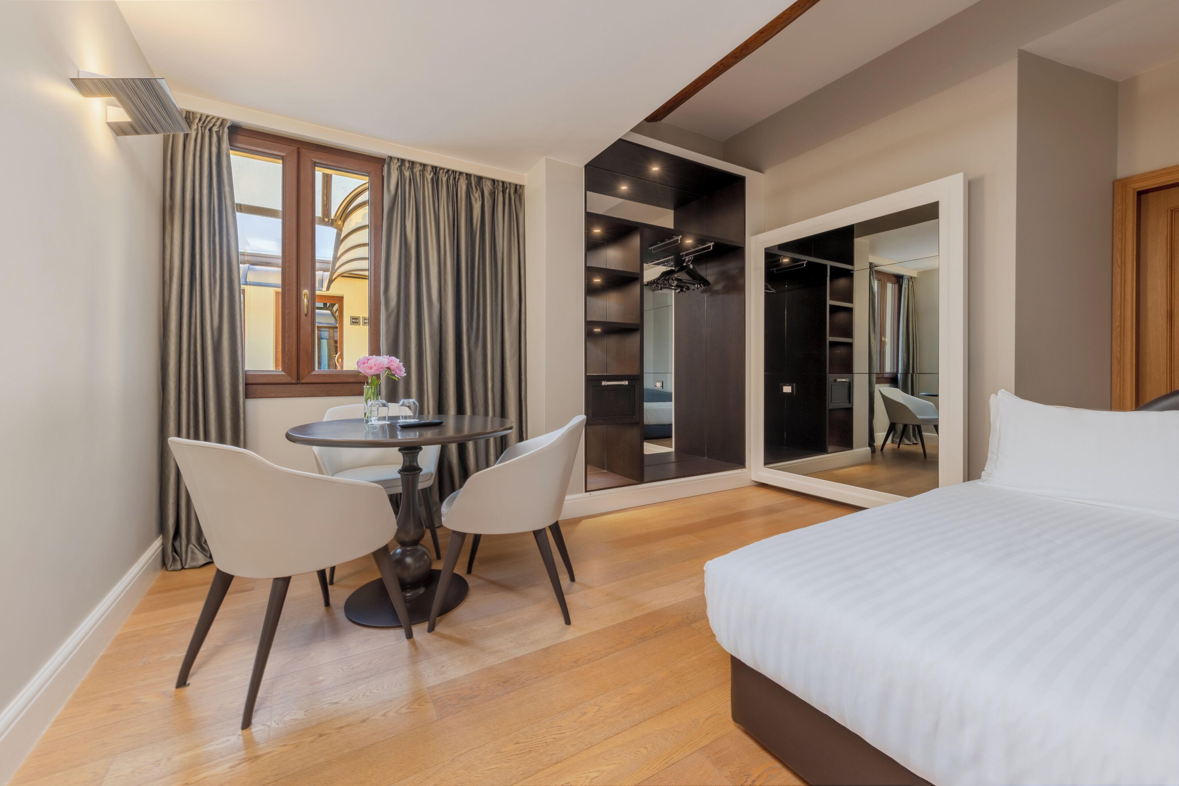 A modern hotel room with a large bed, a dining table with two chairs, a small round table with a vase, and a mirror reflecting the room.