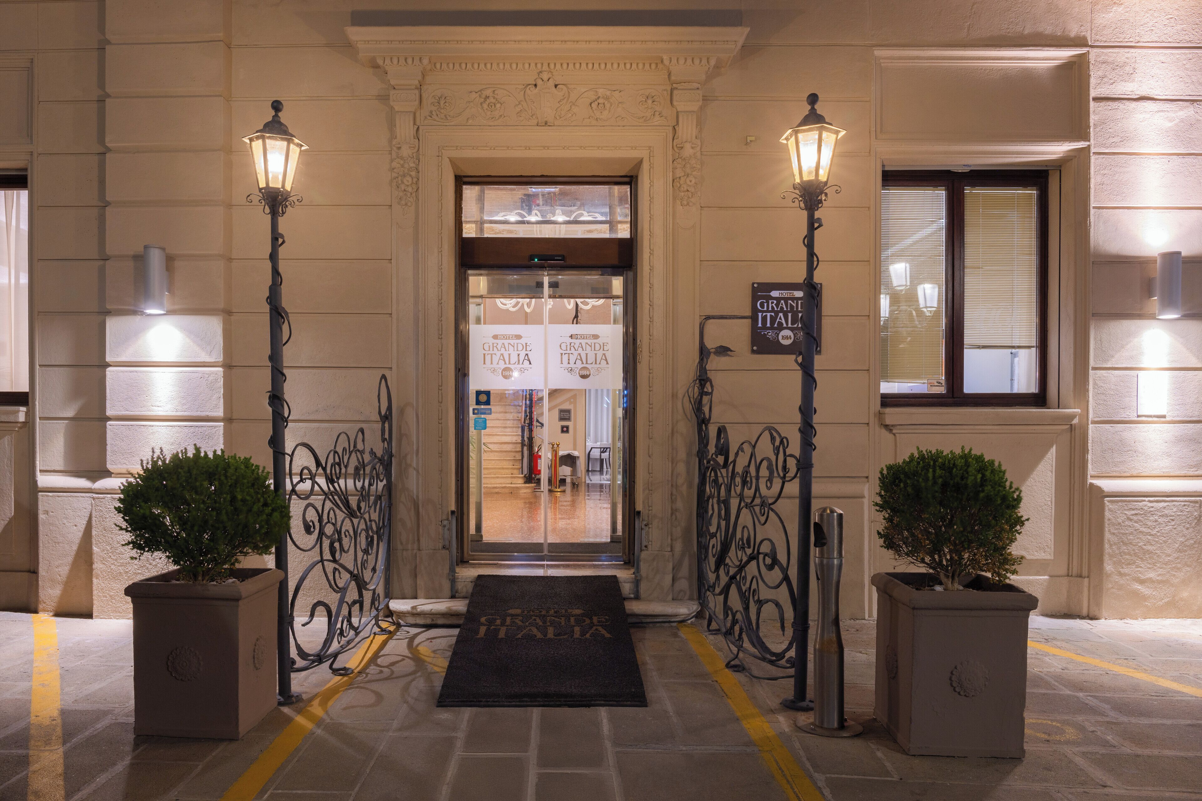 Ein Hotel Eingang mit Doppeltüren, zwei Blumentöpfen, einer Fußmatte und zwei Wandleuchten.