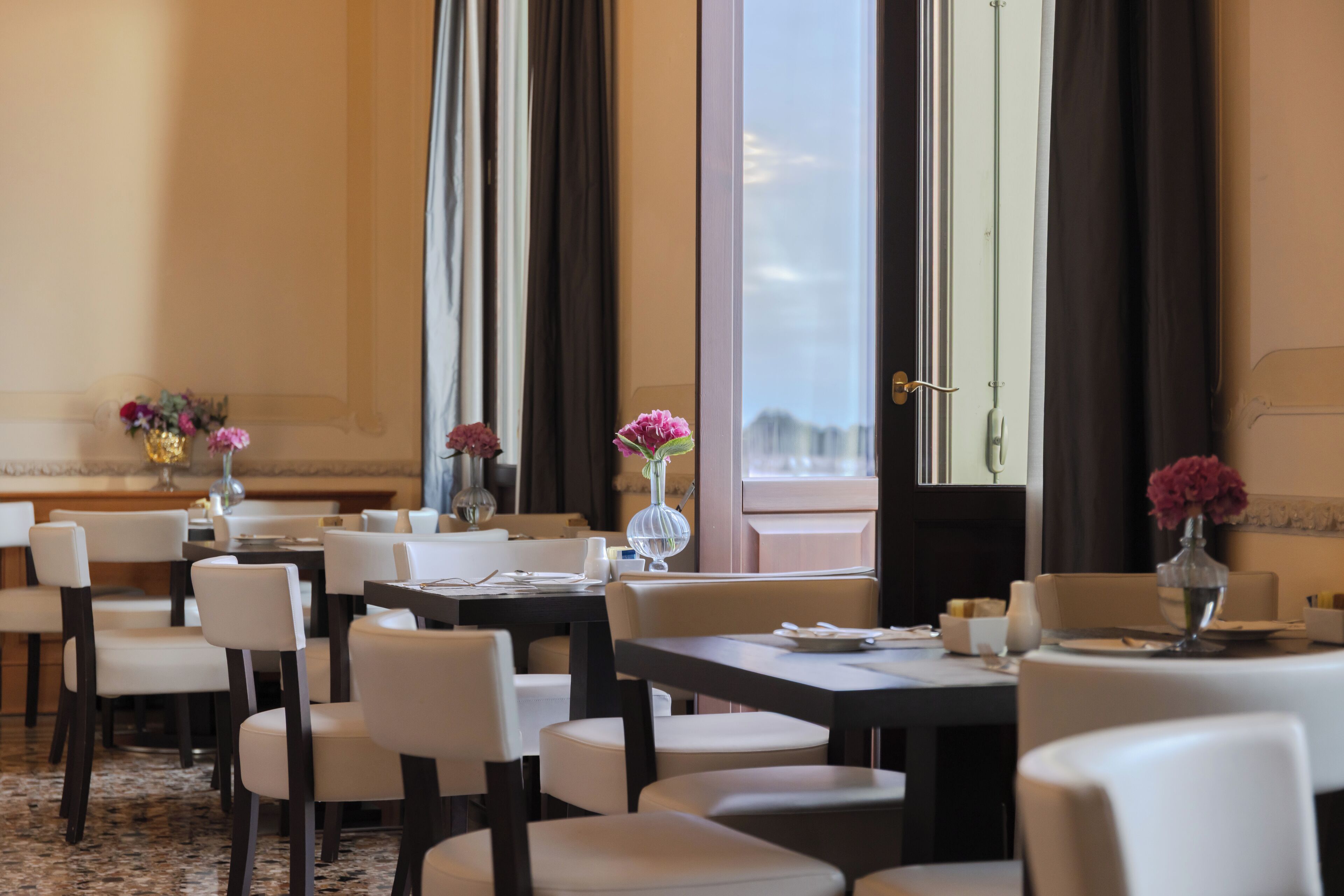 Un espace de restauration d’hôtel comprenant plusieurs tables et chaises, de grandes fenêtres avec des rideaux et des centres de table floraux.