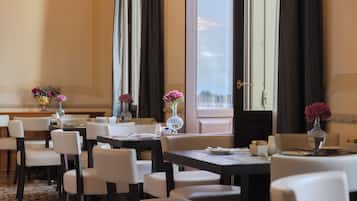 Un comedor de hotel con varias mesas y sillas, ventanas amplias con cortinas y centros de mesa con flores.