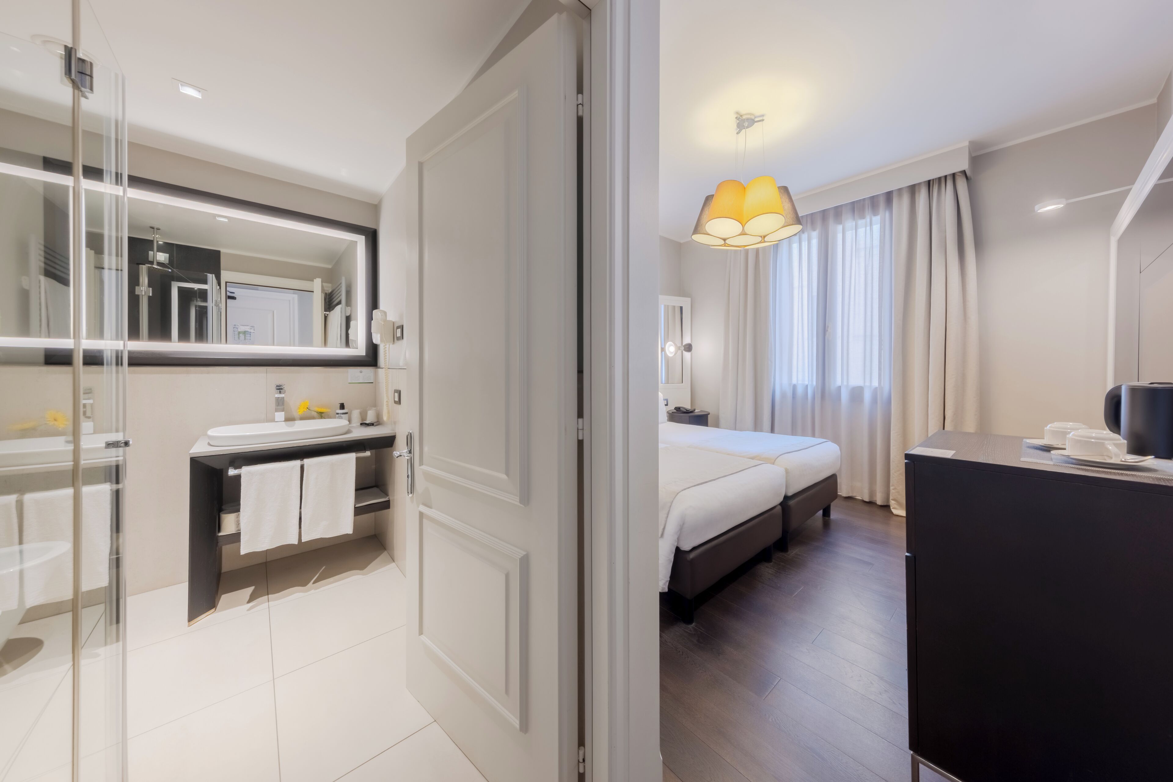 Een moderne hotelkamer met een douchecabine van glas, een grote spiegel, een bed met een witte dekbedovertrek, een nachtkastje en een bureau met een lamp.